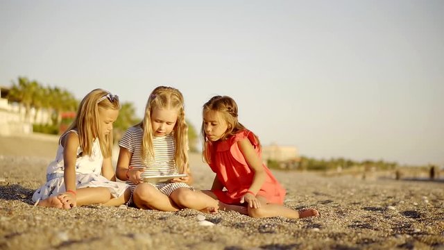 Three little girls are playing on a tablet on beach.