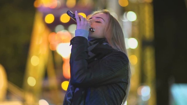 young beautiful woman alone drinks whiskey alcohol from flask. Outdoors at night in an amusement park. Night city background 