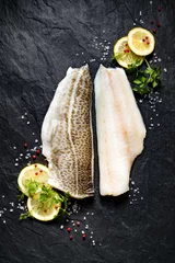 Fotobehang Verse vis, rauwe kabeljauwfilet met toevoeging van kruiden en schijfjes citroen op zwarte stenen ondergrond, bovenaanzicht © zi3000