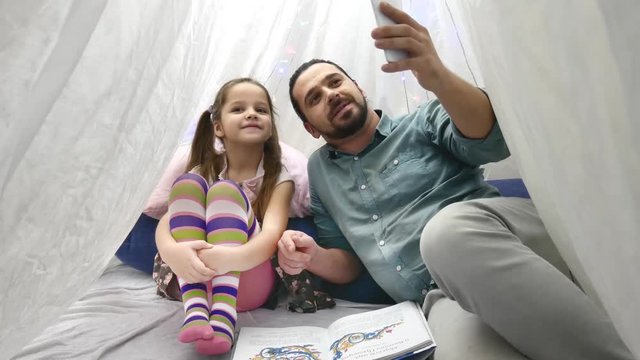 Father with cheerful little daugher playing with smartphone and talking on Skype by mobile