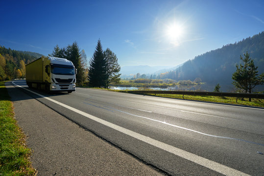 Glowing Sun Over The Valley. White Truck Driving On The Road Around A Lake And Forested Mountains In Autumn.