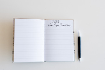 High angle view of lined journal with handwritten 2018 New Year's Resolutions heading and pen on white table (selective focus)