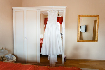 Wedding dress hanging on the wardrobe in the bedroom. The wedding collection. Classic expensive dress for a wedding or celebration in the room of an expensive hotel. Closeup