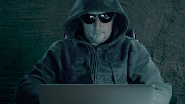 Adult caucasian man in hoody sweatshirt and glasses sits with computer and surf internet late night at concrete background.