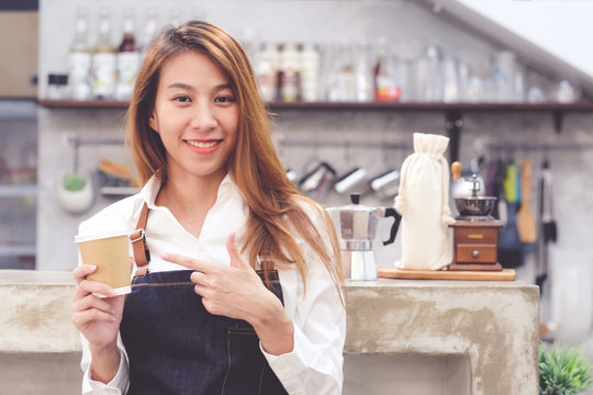Young asia woman barista holding a diaposable coffee cup with smiling face at cafe counter background, small business owner, food and drink industry concept