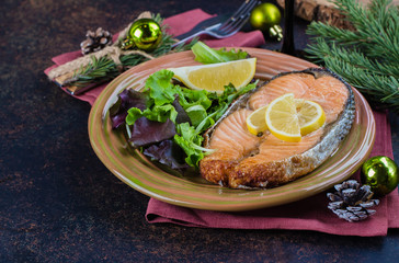Christmas Dinner Table Setting. Delicious salmon steak grilled on plate with lemon and assorted salad on dark stone table background. Top view, copy space