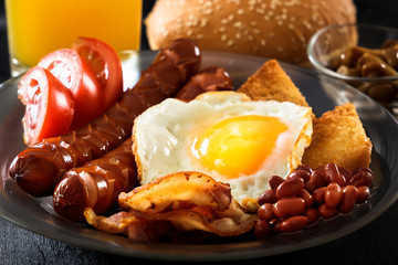 Bacon, grilled sausages, fried egg, beans, beans, toast, tomatoes, mushrooms fresh juice - full English breakfast. Black background. Morning light