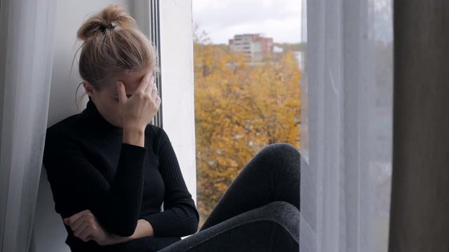 Young depressed and sad woman sitting on the windowsill