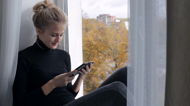 Beautiful woman reads e-book sitting on windowsill