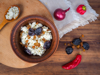 Clay bowl with dairy product cottage cheese, raisins and dried apricots next to the wooden spoon, pepper, paprika and onion on old wood and tablecloth. Template for menu, print design, top view.