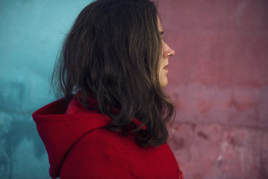 Retrato De Mujer Joven De Perfil De Cabello Largo Castaño Con Piercing En La Nariz Con Chaqueta Roja Con Capucha Sobre Pared De Color Azul Y Roja. Foto Realizada En La Calle.