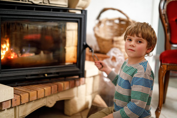 Little cute kid boy sitting by fireplace at home and helping with watching out for fire