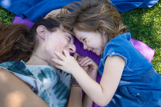 Woman Mother And Four Years Old Blonde Child Laughing Together, Looking With Complicity, Lying On Towels In The Green Grass Of Park
