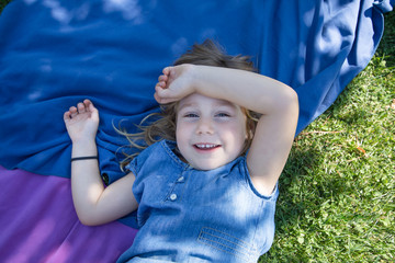 four years old blonde girl lying on towels in the green grass of park with hand in head looking and smiling
