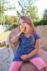 four years old blonde girl with blue denim dress sitting in public park, biting orange or yellow ice lolly or popsicle
