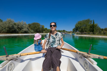 woman mother paddling in boat, smiling happy, next to four years old blonde girl, at park lake 
