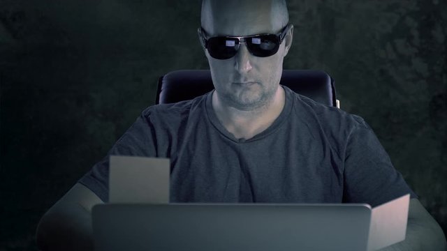 A man in glasses and a T-shirt at night at a table with a laptop. The hacker completed the task and removed the stickers of the note from the laptop screen.
