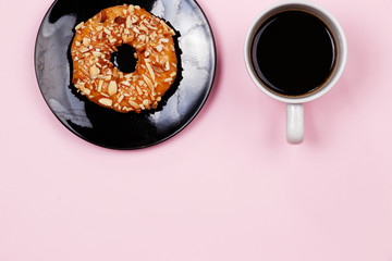 Almond caramel donut and black coffee with copy space. Cup of black coffee with donut on a black plate on pink background, top view, flat lay. Delicious breakfast.