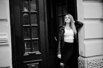 Elegant blonde girl wear on black leather jacket posing at streets of town background old doors.