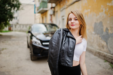 Naklejka na ściany i meble Elegant blonde girl wear on black leather jacket posing at streets of town background luxury car.