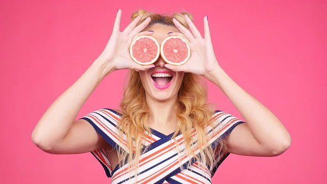 Young happy woman is holding slices of grapefruits to her eyes on pink background