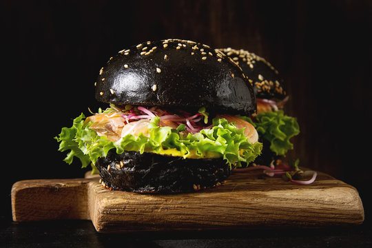 Black burger with salmon fish, lettuce, mustard. Dark background. Fast food.