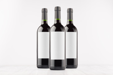 Three dark wine bottles with blank white labels on white wooden board, mock up. Template for advertising, design, branding identity.
