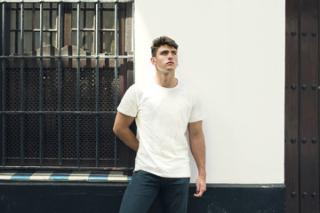 Retrato de hombre joven con camiseta blanca apoyado en la pared de una casa en una calle típica...