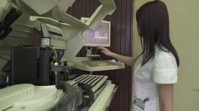 Close up of young pretty female technician in white coat during typing and loading parameters on  computer touch screen at laboratory room, automated machine, blood and urine sample test, crane shot