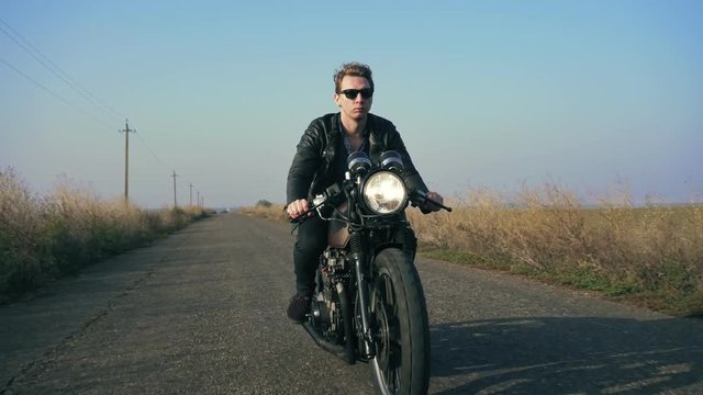 Closeup view of a stylish cool young man in sunglasses and leather jacket riding motorcycle on a road on a sunny day. Then the camera moves forward and the man visibly shrinking into the distance