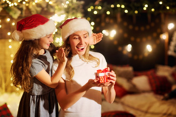 Mother and daughter in Santa hats give presents in a room with a fireplace and a Christmas tree on Christmas.