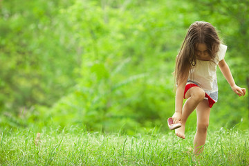 Naklejka na ściany i meble After rain concept. Stylish baby girl with long light brown hair in trendy vintage red and white dress standing in the park and trying to put on a shoe. Sunny weather. Copy-space