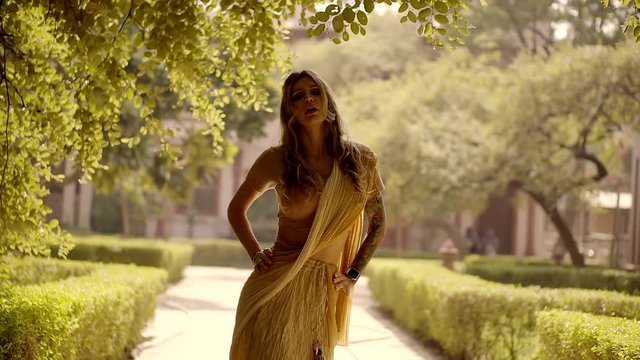 sensual adult blonde is dancing oriental dance in a garden, moving hips and hands, sun is shining