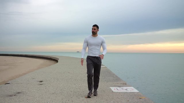 Male runner is walking along beautiful seashore while listening to the music with headphones. Ready to workout.