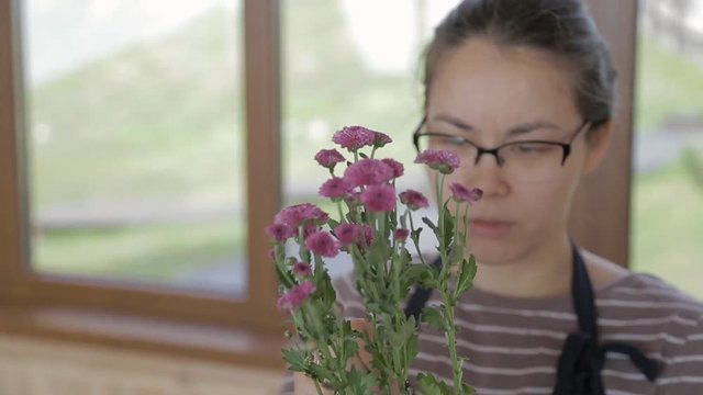 Young woman is holding bouquet of lilac chrysanthemums in hands indoors. Female in glasses has annual plant with green leaves and small fluffy flowers. Attractive brunette stands by window, admires
