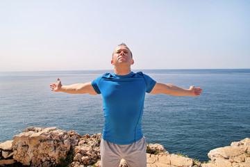 Athletic fit runner doing stretching exercise, preparing for morning workout on a seashore. A handsome fitness athlete doing a stretching routine in the sun. Blue sky and the sea in background.