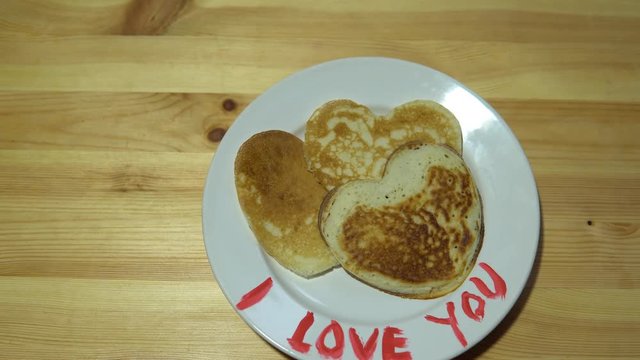 Hands take pancakes in the shape of a heart with a white plate with a red inscription I love you.