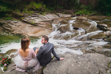 Groom in grey suit and bride in a light dress pose on the rocks before a mountain river