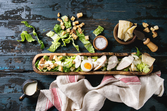 Classic Caesar salad with grilled chicken breast and half of egg in olive wood bowl. Served on kitchen towel with ingredients above over old dark blue wooden background. Top view, space. Rustic style