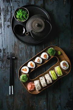 Homemade sushi rolls set with salmon, sesame seeds and avocado serving in wood plate with pink pickled ginger, soy sauce, wasabi, seaweed salad, chopsticks over dark wooden background. Top view, space