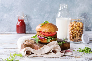 Homemade burger in classic bun with tomato sauce, arugula, meat, cheese, onion on wood serving board with bottle of ketchup, milk, roast chickpeas over white wooden plank table.