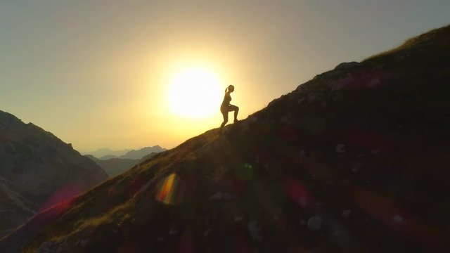 AERIAL SILHOUETTE: Flying over young woman hiker as she climbs up sloping terrain on her way to summit. Woman trying to reach mountain peak before sundown. Lady climbing uphill in beautiful scenery.