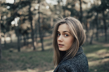 Gentle and pretty young woman with long blonde hair looking at camera over shoulder . Model posing at camera in coat in park outdoor