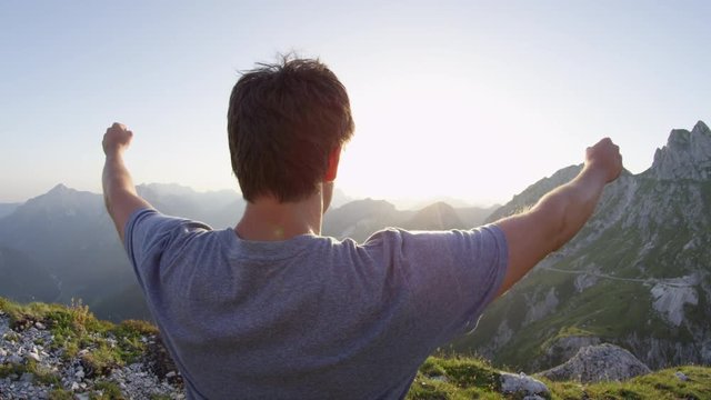CLOSE UP LENS FLARE: Joyful young man standing on mountaintop, outstretching arms victoriously at stunning summer sunset. Happy male hiker celebrating successful mountain ascent and enjoying the view.