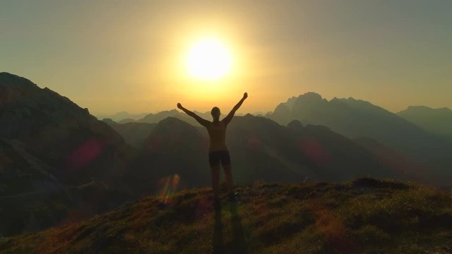 AERIAL Flying over hiker woman walking up the mountain slope and raising her arms victoriously at golden summer sunset. Silhouette of girl hiking high rocky mountains and celebrating successful ascent