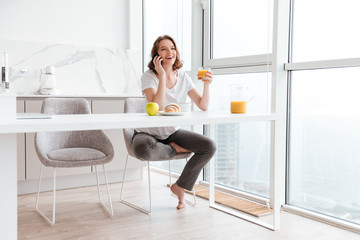 Fototapeta na wymiar Young attractive woman in casual wear speaking on mobile phone and holding glass of orange juice while sitting at kitchen table