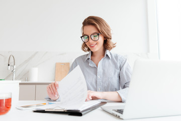 Photo of cheerful attractive woman in glasses reading new contract while working in the kitchen