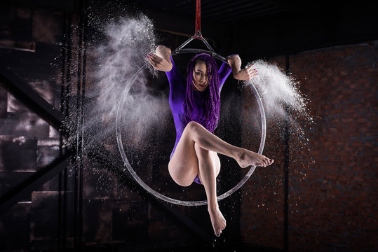 A young, slender girl in a lavender leotard and lilac dreads, doing gymnastics on the air ring, the Hoop and throws the hands of the white powder in the form of a cloud.