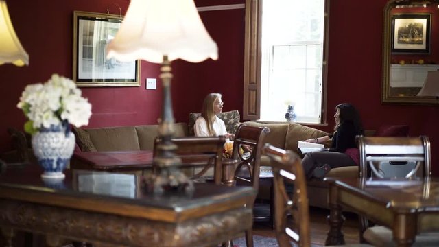 Two women sit on couches and talk in a grand room in a historic inn with red walls and large windows.