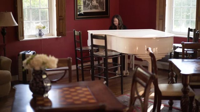 A woman plays a white grand piano in a red colonial room in a historic inn with large windows.